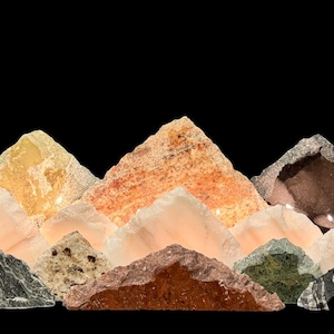 May include: A collection of various natural stones and minerals arranged to resemble a mountain range. The pieces feature diverse textures and colors, including shades of orange, white, grey, brown, green, and black, with some translucent elements.