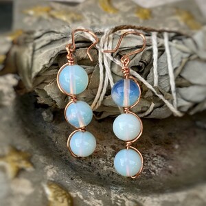 May include: A pair of dangle earrings featuring three round, translucent, pale blue opal-like beads each, wrapped in copper wire. The earrings hang from copper ear wires, showcasing a smooth, polished finish on the spherical stones and a metallic copper frame.