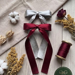 Könnte beinhalten: Silberne und bordeauxfarbene Samtbandschleifen mit passenden Bandschwänzen, präsentiert mit einer Spule bordeauxfarbenen Fadens, Trockenblumen und Baumwollkapseln. Ein grüner und goldener "Fine Ribbon"-Aufkleber ist ebenfalls vorhanden.