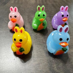 May include: Five small rubber duck toys shaped like bunnies, each holding a carrot. The ducks are pink, green, purple, blue, and yellow.