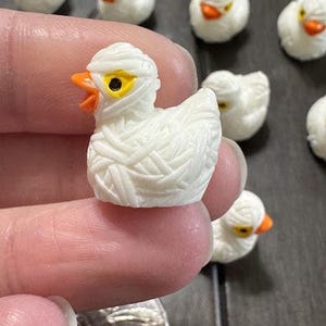 May include: A collection of small white ceramic duck figurines. One duck is wrapped in white gauze, resembling a mummy. The ducks have orange beaks and yellow eyes.