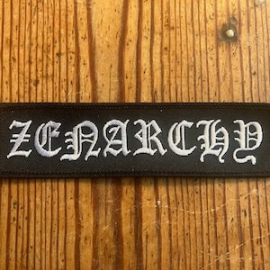 Pode incluir: Um remendo de tecido preto com letras bordadas brancas que dizem "ZENARCHY".