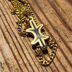 May include: A gold chain necklace with a black and gold pendant featuring a stylized symbol. The pendant is in the shape of a cross with a circle in the center.