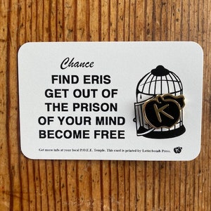 May include: A white card with black text that reads "Chance Find Eris Get Out Of The Prison Of Your Mind Become Free". The card has a black and white illustration of a birdcage with a black circle inside that has a gold letter "K" on it. The card also has a small black "K" in the bottom right corner.