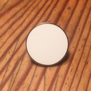 May include: A white enamel pin with a gold border. The pin is round and has a smooth surface.