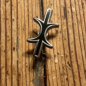 May include: A silver and black enamel pin with a stylized design of a crescent moon and a vertical line.