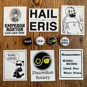Op de afbeelding: Een verzameling stickers en knoppen met de tekst "Hail Eris" en andere gerelateerde zinnen. De stickers zijn zwart-wit en hebben verschillende ontwerpen, waaronder een cartoonkat, een yin-yang symbool en een figuur met een vredesteken. De knoppen zijn rond en hebben verschillende kleuren en ontwerpen.