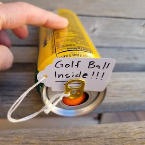 Può includere: Una lattina di alluminio gialla con un'etichetta bianca attaccata alla linguetta di apertura. L'etichetta dice "Golf Ball Inside!!!". È possibile vedere una palla da golf all'interno della lattina.
