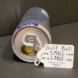 May include: A silver can with a yellow golf ball inside. The can has a pull tab and a label that says "Golf Ball in a SMALL can in a LARGE can".