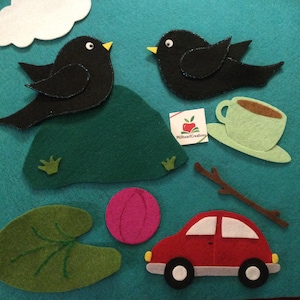 May include: A felt craft scene with two black birds, a green hill, a white cloud, a pink ball, a red car, a green leaf, a cup of coffee, and a brown stick on a teal background. The scene is likely for children.