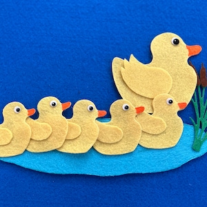 Peut inclure: Une création en feutre représentant un canard jaune avec cinq canetons sur un étang bleu avec des roseaux verts. Les canetons sont alignés en rang derrière la cane.