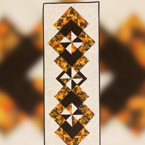 May include: A rectangular quilt runner featuring a repeating geometric pattern of diamonds and triangles in shades of brown, orange, and white. The text "SPIN SET RUNNER" and "DESIGNED BY MAD B'S QUILT & SEW" are at the top. The Mad B's logo and website are at the bottom.