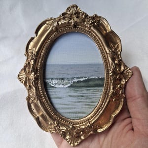 Könnte beinhalten: Ein ovaler Bilderrahmen mit einem verzierten, metallisch goldenen Finish, das komplizierte Blumen- und Schnörkeldetails aufweist. Der Rahmen enthält ein kleines Gemälde einer Meereslandschaft mit einem ruhigen Ozean und einer weißen Welle.