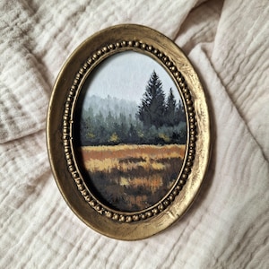 Könnte beinhalten: Ein ovales Gemälde in einem goldfarbenen Rahmen mit perlenartigem Innenrand. Das Kunstwerk zeigt eine Landschaft mit goldbraunem Gras im Vordergrund, dunkelgrünen Kiefern im Mittelgrund und einem nebligen, hellgrauen Himmel im Hintergrund.