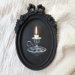 Könnte beinhalten: Ein ovaler schwarzer Rahmen mit einer dekorativen Schleife und floralen Reliefdetails umgibt ein Gemälde einer brennenden Kerze in einem silbernen Kerzenhalter. Die Kerze hat eine helle Flamme und der Kerzenhalter einen kleinen Griff.