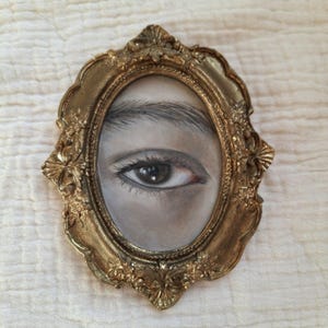Könnte beinhalten: Ovaler, antikgoldfarbener Rahmen mit aufwendigem Design. Der Rahmen umgibt eine Graustufenzeichnung eines menschlichen Auges und einer Augenbraue. Das Auge ist braun mit sichtbaren Wimpern und einer detaillierten Iris.