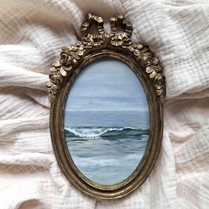 Könnte beinhalten: Ein ovales Gemälde einer Meereslandschaft mit einer brechenden Welle, ruhigem blauem Wasser und einem blassen Himmel mit kleinen Vögeln, eingefasst in einen verzierten, metallisch goldfarbenen Rahmen mit skulpturalen Blumen- und Schleifendetails.