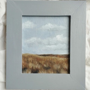 Könnte beinhalten: Ein gerahmtes Gemälde einer Landschaft mit einem Feld aus braunem Gras und einem blauen Himmel mit weißen Wolken.