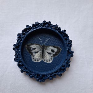 Könnte beinhalten: Ein kleiner, runder, marineblauer dekorativer Rahmen mit einem verzierten, strukturierten Rand. Im Inneren des Rahmens befindet sich ein Gemälde eines Schmetterlings mit weißen und grauen Flügeln. Der Schmetterling ist auf einem marineblauen Hintergrund zentriert.