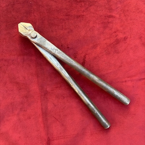 May include: A pair of metal end cutting pliers with a weathered, matte gray finish. The tool features two long, straight handles connected by a central pivot pin, leading to a sharp, angled cutting head designed for gripping and cutting wire.