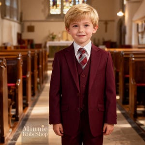 May include: A young person wearing a burgundy three-piece suit with a white collared shirt and a plaid tie. The suit includes a jacket, vest, and pants. The background is a church interior with wooden pews.
