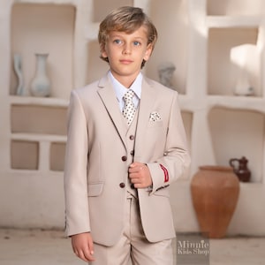 May include: A beige three-piece suit for boys, featuring a jacket, vest, and pants. The suit includes a white dress shirt and a patterned tie. The suit is well-tailored and suitable for formal occasions. The suit is from Minnie Kids Shop.