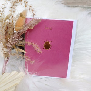 Könnte beinhalten: Eine rosa Karte mit einem goldenen Käfer und dem Wort "love" in goldenen Lettern.