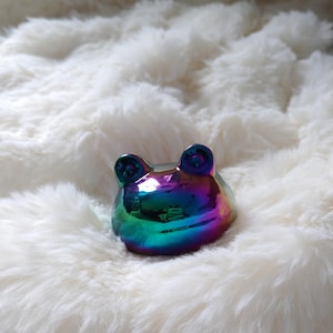Op de afbeelding: Een kleine, iriserende kikkerfiguur met een regenboogafwerking. De keramische kikker heeft grote, ronde ogen en staat op een zacht, wit oppervlak. De kleuren van de kikker verschuiven van blauw naar groen naar paars.