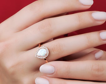 Puede incluir: Un anillo de oro rosa con una piedra de ópalo en forma de lágrima rodeada por un halo de piedras pequeñas transparentes.