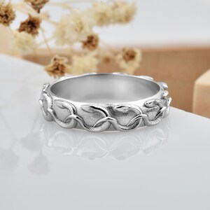 May include: A silver-toned metal ring featuring a continuous, embossed design of intertwined snakes wrapping around the entire band. The surface of the ring has a textured, matte finish between the raised, polished snake figures.