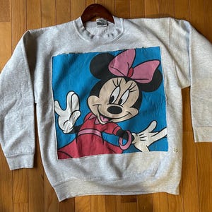 Puede incluir: Sudadera gris claro con un gran gr&aacute;fico de Minnie Mouse. Minnie se representa con un lazo rosa, un vestido rojo y guantes blancos sobre un fondo azul. La sudadera tiene mangas largas y cuello redondo.