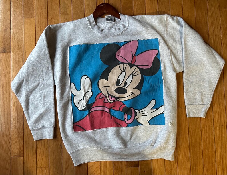 Puede incluir: Sudadera gris claro con un gran gr&aacute;fico de Minnie Mouse. Minnie se representa con un lazo rosa, un vestido rojo y guantes blancos sobre un fondo azul. La sudadera tiene mangas largas y cuello redondo.
