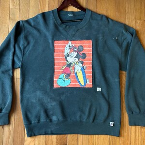 May include: Dark green sweatshirt featuring a graphic of Mickey Mouse on a skateboard, set against a red and orange striped background. The sweatshirt has a crew neck and long sleeves. A small white tag is visible on the lower right.