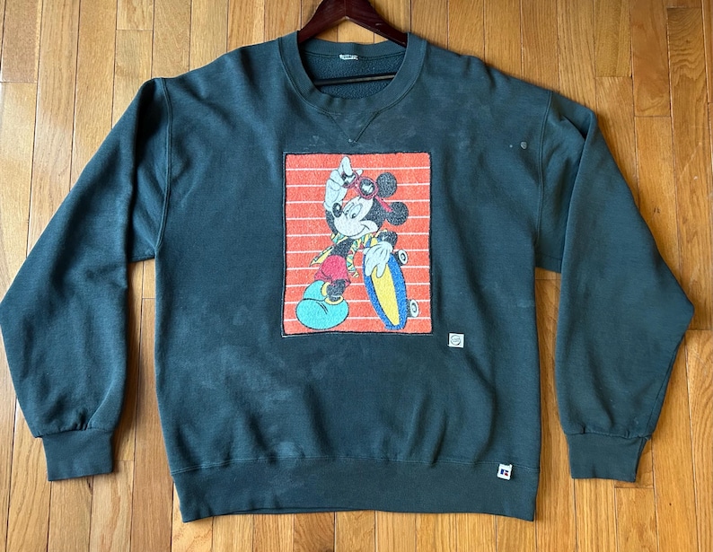 May include: Dark green sweatshirt featuring a graphic of Mickey Mouse on a skateboard, set against a red and orange striped background. The sweatshirt has a crew neck and long sleeves. A small white tag is visible on the lower right.