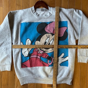 Puede incluir: Una sudadera gris de cuello redondo con un gran gr&aacute;fico de Minnie Mouse. Minnie se representa con un lazo rosa, un vestido rojo y guantes blancos sobre un fondo azul. La sudadera se muestra en una percha de madera.