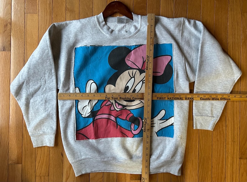 Puede incluir: Una sudadera gris de cuello redondo con un gran gr&aacute;fico de Minnie Mouse. Minnie se representa con un lazo rosa, un vestido rojo y guantes blancos sobre un fondo azul. La sudadera se muestra en una percha de madera.