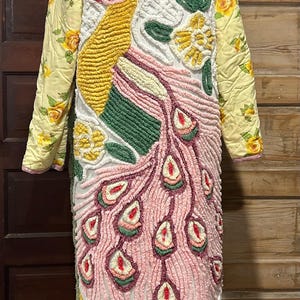 May include: A long, quilted coat with a vibrant peacock design. The coat features a white base with a textured pattern, a yellow body, and sleeves with a floral print. The peacock's tail is pink with red and green accents.