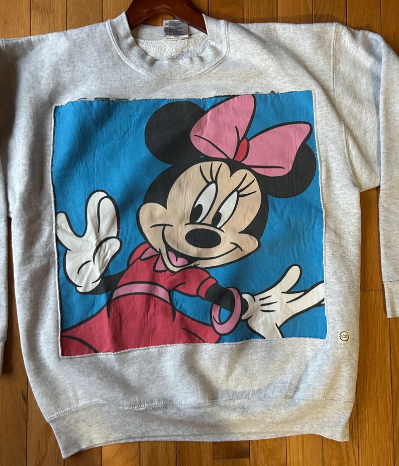 Puede incluir: Sudadera gris con un gran gr&aacute;fico de Minnie Mouse. El gr&aacute;fico muestra a Minnie Mouse con un vestido rojo y un lazo rosa sobre un fondo azul. La sudadera tiene cuello redondo y mangas largas.