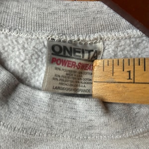 Puede incluir: Primer plano de una sudadera gris con una etiqueta que dice "ONEITA POWER-SWEAT". La etiqueta tambi&eacute;n indica que la sudadera est&aacute; hecha en los EE. UU. y es una mezcla de poli&eacute;ster y algod&oacute;n. El tama&ntilde;o es "LARGE/GRAND/GRANDE."