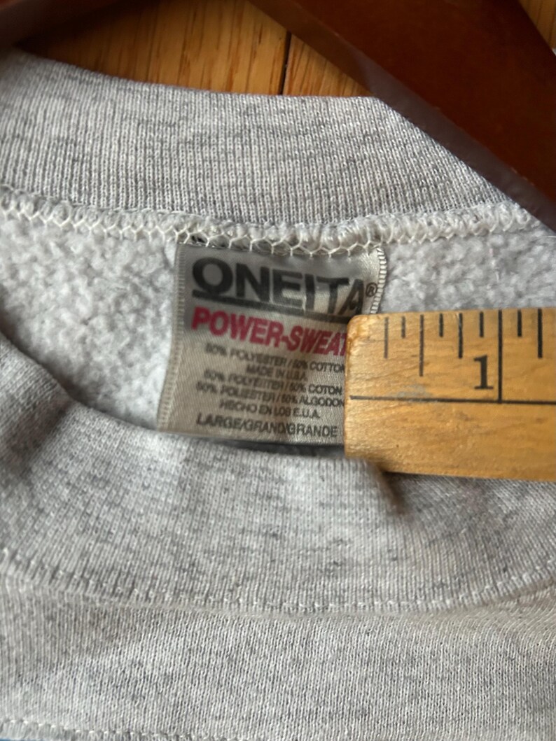 Puede incluir: Primer plano de una sudadera gris con una etiqueta que dice "ONEITA POWER-SWEAT". La etiqueta tambi&eacute;n indica que la sudadera est&aacute; hecha en los EE. UU. y es una mezcla de poli&eacute;ster y algod&oacute;n. El tama&ntilde;o es "LARGE/GRAND/GRANDE."