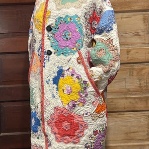 Könnte beinhalten: Ein langärmeliger Mantel aus weißem Steppstoff mit einem mehrfarbigen Patchwork-Design aus hexagonalen Blumenmustern in Rot, Gelb, Blau, Lila und Grün. Der Mantel verfügt über einen hohen Kragen, zwei dunkle Knöpfe und aufgesetzte Taschen.