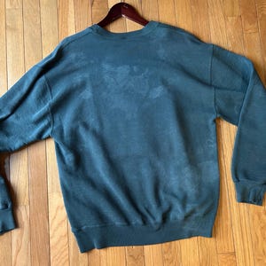 May include: A dark teal sweatshirt with long sleeves, a crew neck, and ribbed cuffs and hem. The sweatshirt is hanging on a wooden hanger and is displayed on a wooden floor.