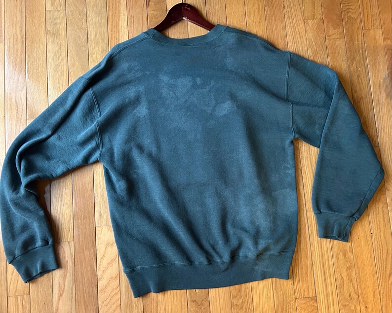 May include: A dark teal sweatshirt with long sleeves, a crew neck, and ribbed cuffs and hem. The sweatshirt is hanging on a wooden hanger and is displayed on a wooden floor.