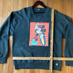 May include: Dark green sweatshirt featuring a graphic of Mickey Mouse skateboarding. The graphic is set against a red and white striped background. The sweatshirt has long sleeves and a crew neck.