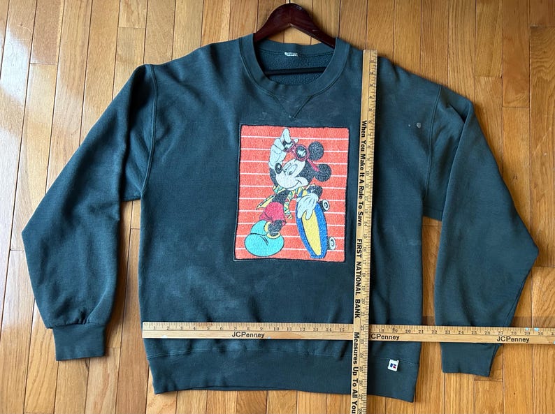 May include: Dark green sweatshirt featuring a graphic of Mickey Mouse skateboarding. The graphic is set against a red and white striped background. The sweatshirt has long sleeves and a crew neck.