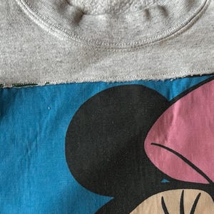 Puede incluir: Una sudadera gris con un parche azul con un gr&aacute;fico de Minnie Mouse. El gr&aacute;fico incluye una cabeza negra, un lazo rosa y una cara beige. La sudadera tiene cuello redondo.