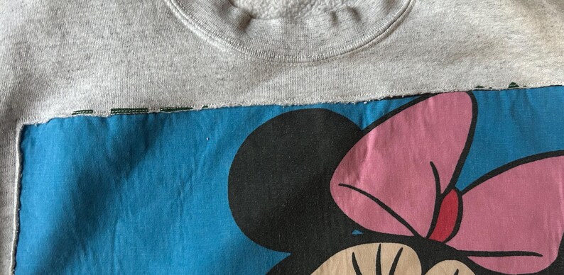 Puede incluir: Una sudadera gris con un parche azul con un gr&aacute;fico de Minnie Mouse. El gr&aacute;fico incluye una cabeza negra, un lazo rosa y una cara beige. La sudadera tiene cuello redondo.