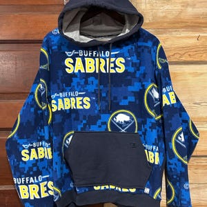 May include: A navy blue and royal blue fleece hoodie featuring a repeating Buffalo Sabres logo and text pattern. The garment includes a solid navy blue front kangaroo pocket with a small Champion logo, a drawstring hood, and ribbed cuffs.