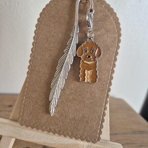 May include: A silver bookmark featuring a feather design and a charm of a brown poodle dog. The bookmark is attached to a brown felt tag with a scalloped edge. The tag is displayed on a small wooden easel.