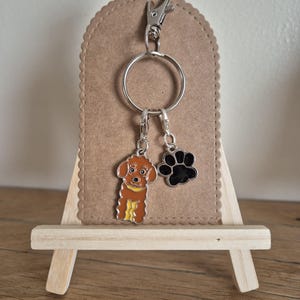 May include: A silver keyring with two charms: a brown dog and a black paw print. The dog charm has yellow detailing. The keyring is attached to a brown, scalloped-edge backing.